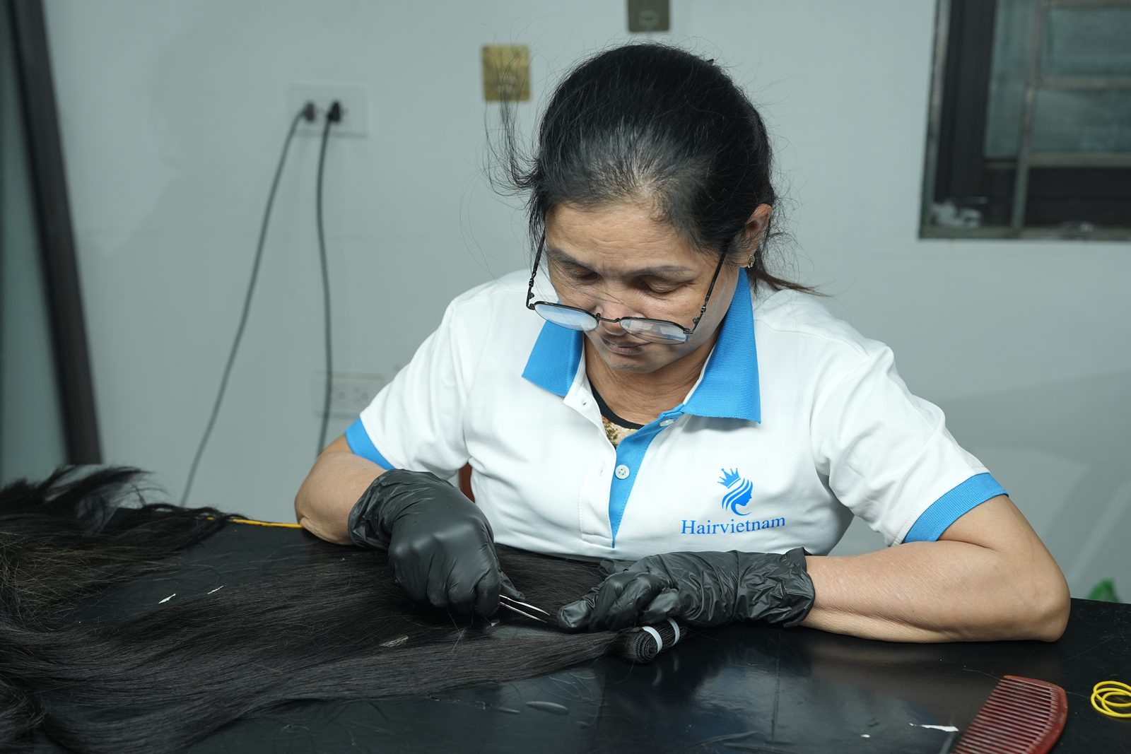 A woman in gloves picks through long dark hair on a black table with tweezers