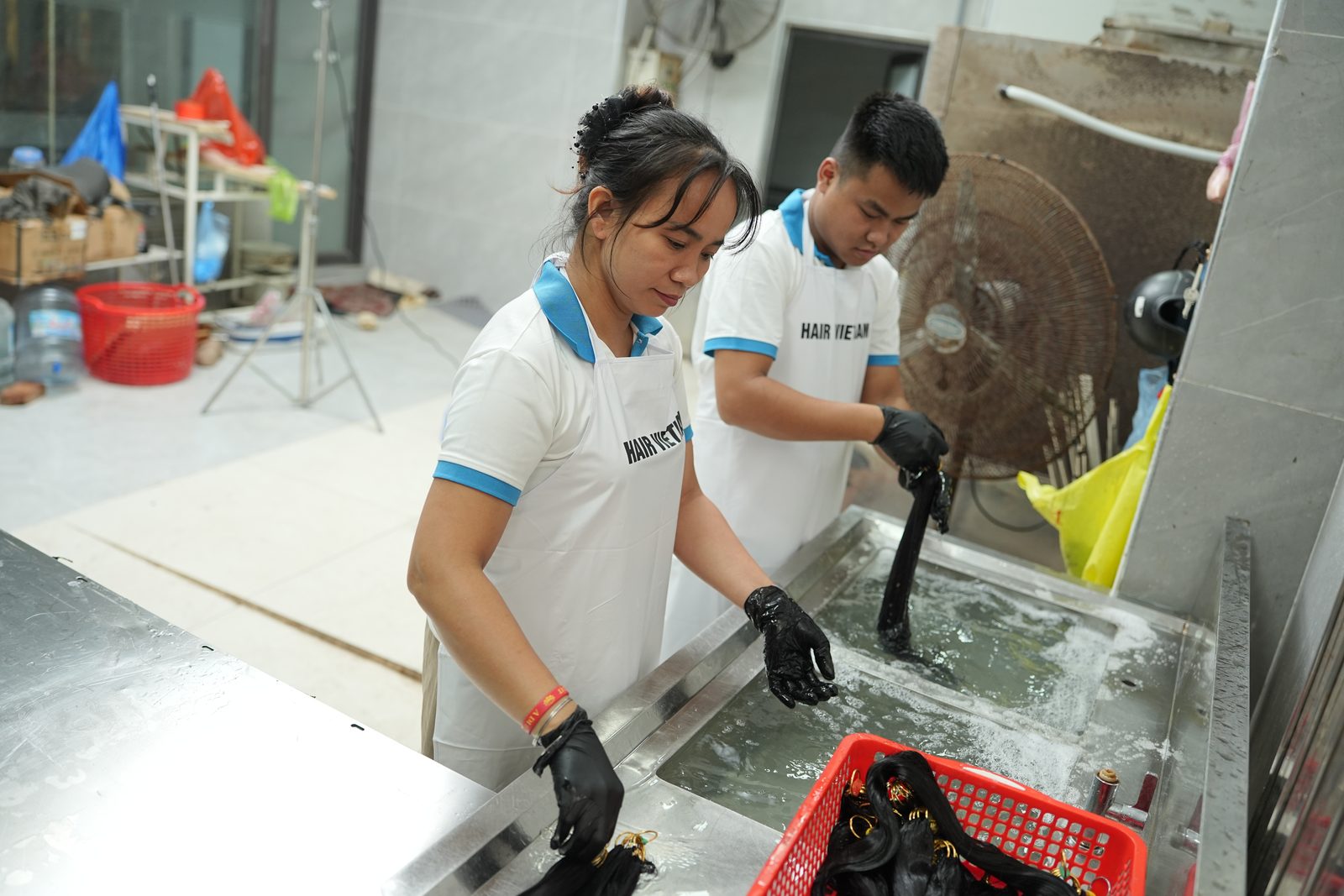 Two staff in aprons rinse dark hair bundles in a stainless-steel sink beside a red crate of bundles